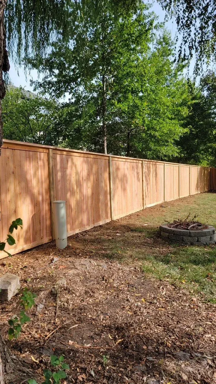 Wooden fence bordering a backyard with a fire pit and trees in the background.
