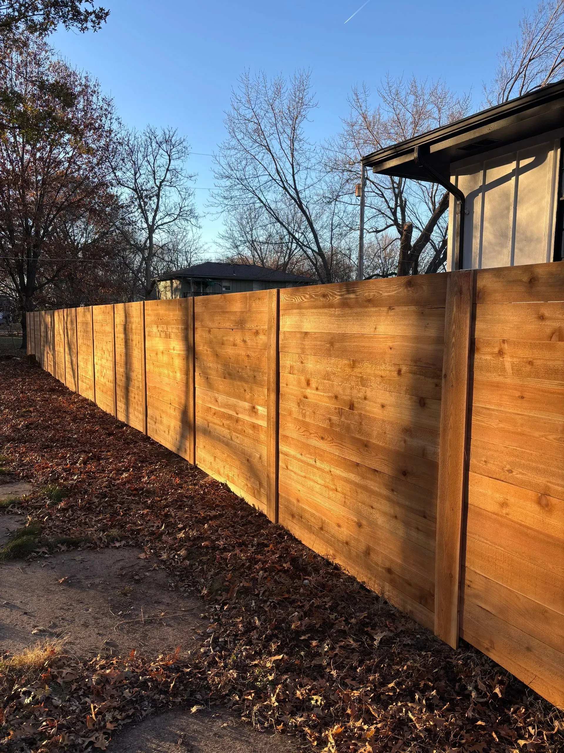 Wooden fence in sunlight with fallen leaves. Side of a building visible. Blue sky.