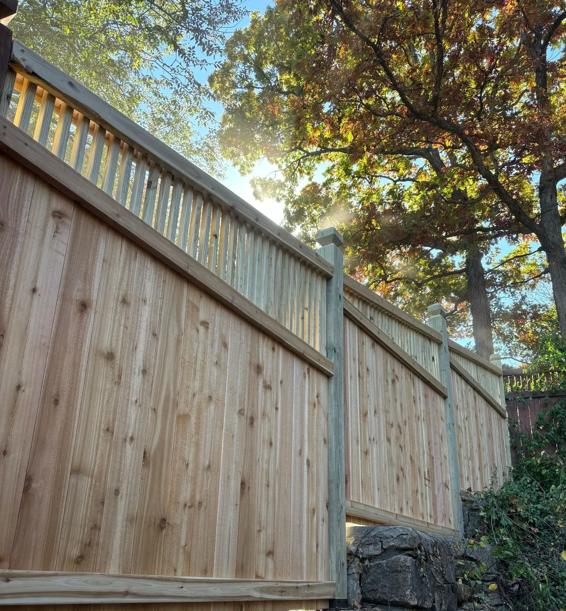 Wooden privacy fence on a stone wall, sunlight through trees.