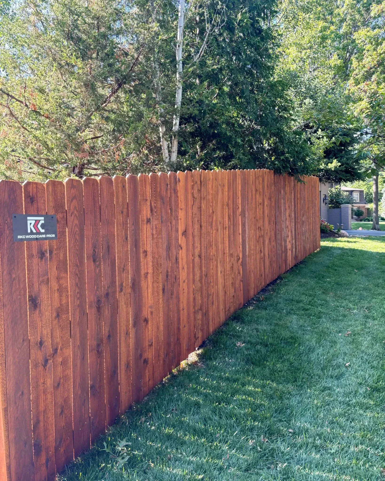 Wooden fence stained reddish-brown, bordering green lawn, trees in background, and a sign on fence.