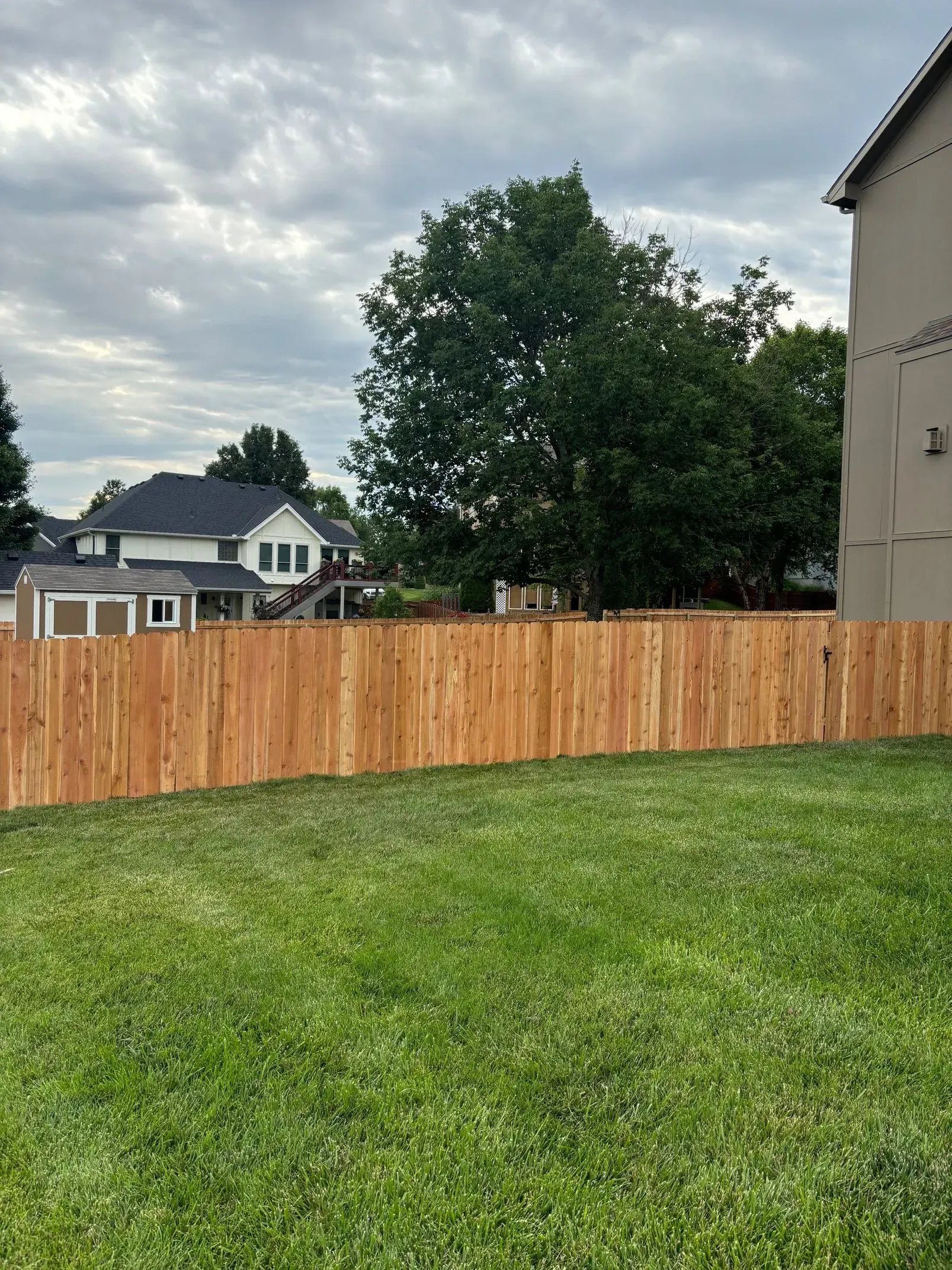Wooden fence in a grassy yard, with houses and trees in the background under a cloudy sky.