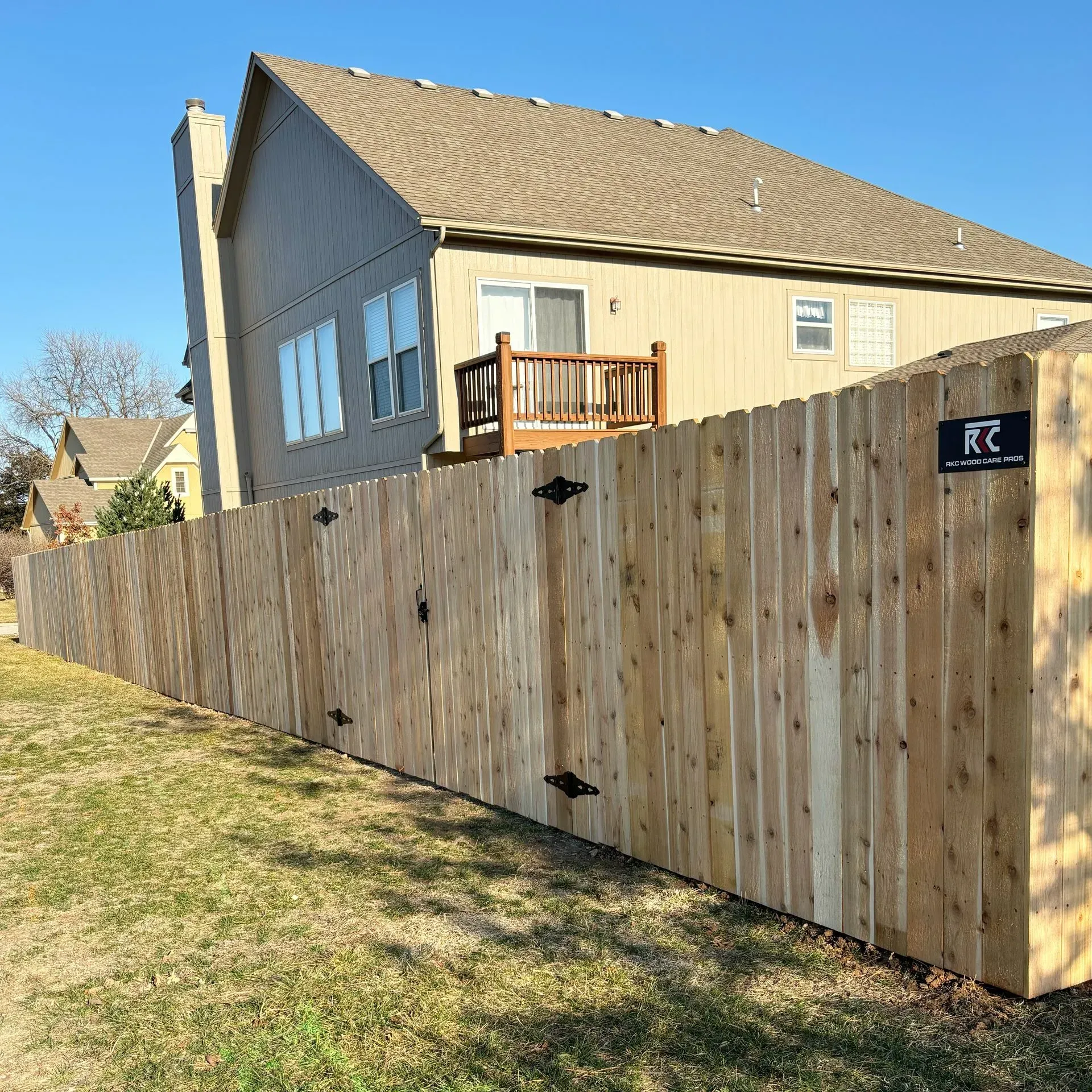Wooden privacy fence along a suburban home; light brown with black hardware, green grass, and blue sky.