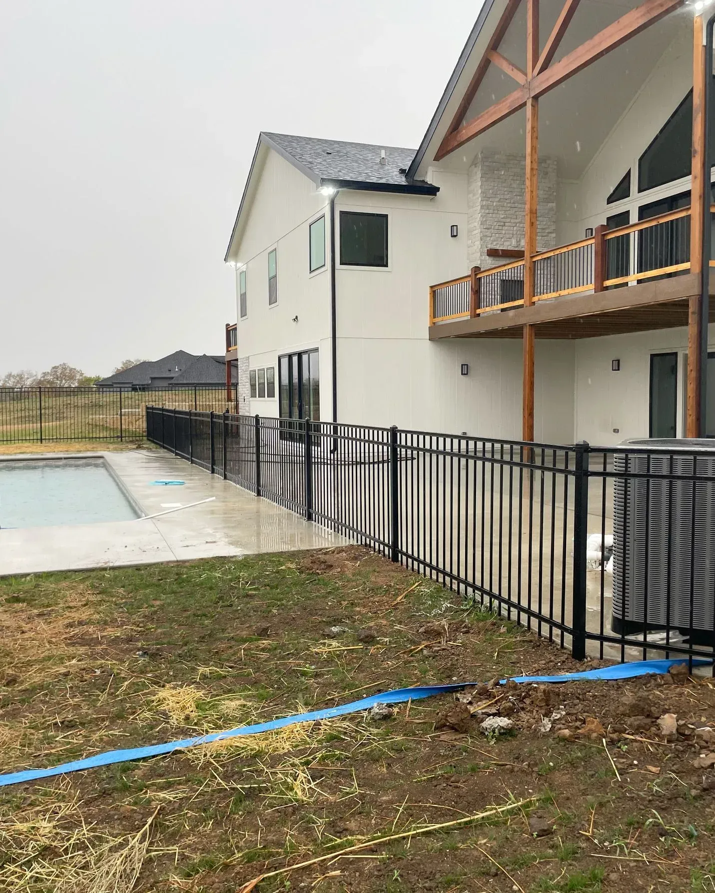 Black metal fence surrounds a white house with a balcony and a pool on a rainy day.
