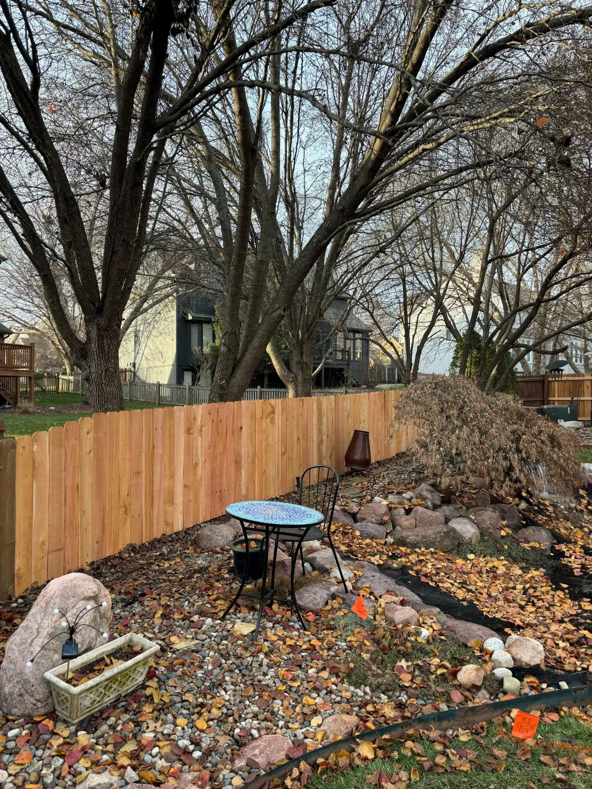 Wooden fence in a backyard with a small table and chairs, large rock pile and fallen leaves.