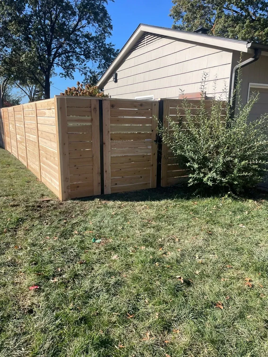 Wooden fence with a gate in a grassy yard next to a house with beige siding.