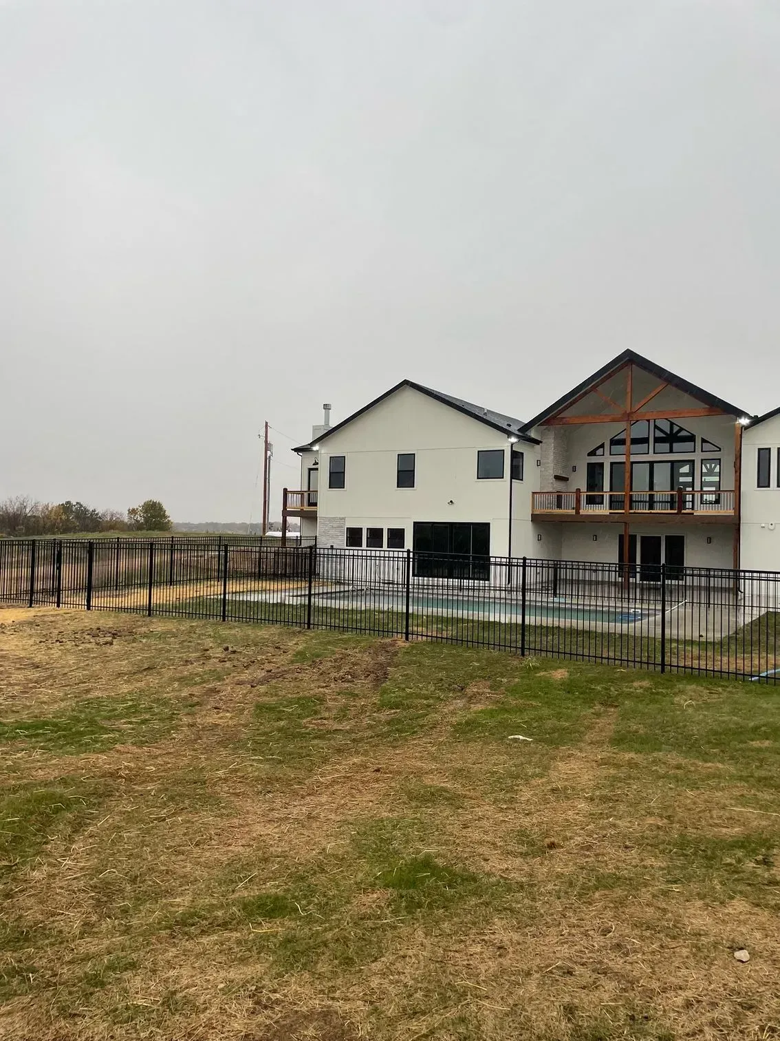 Modern house with pool, black fence, and brown grass yard under a cloudy sky.