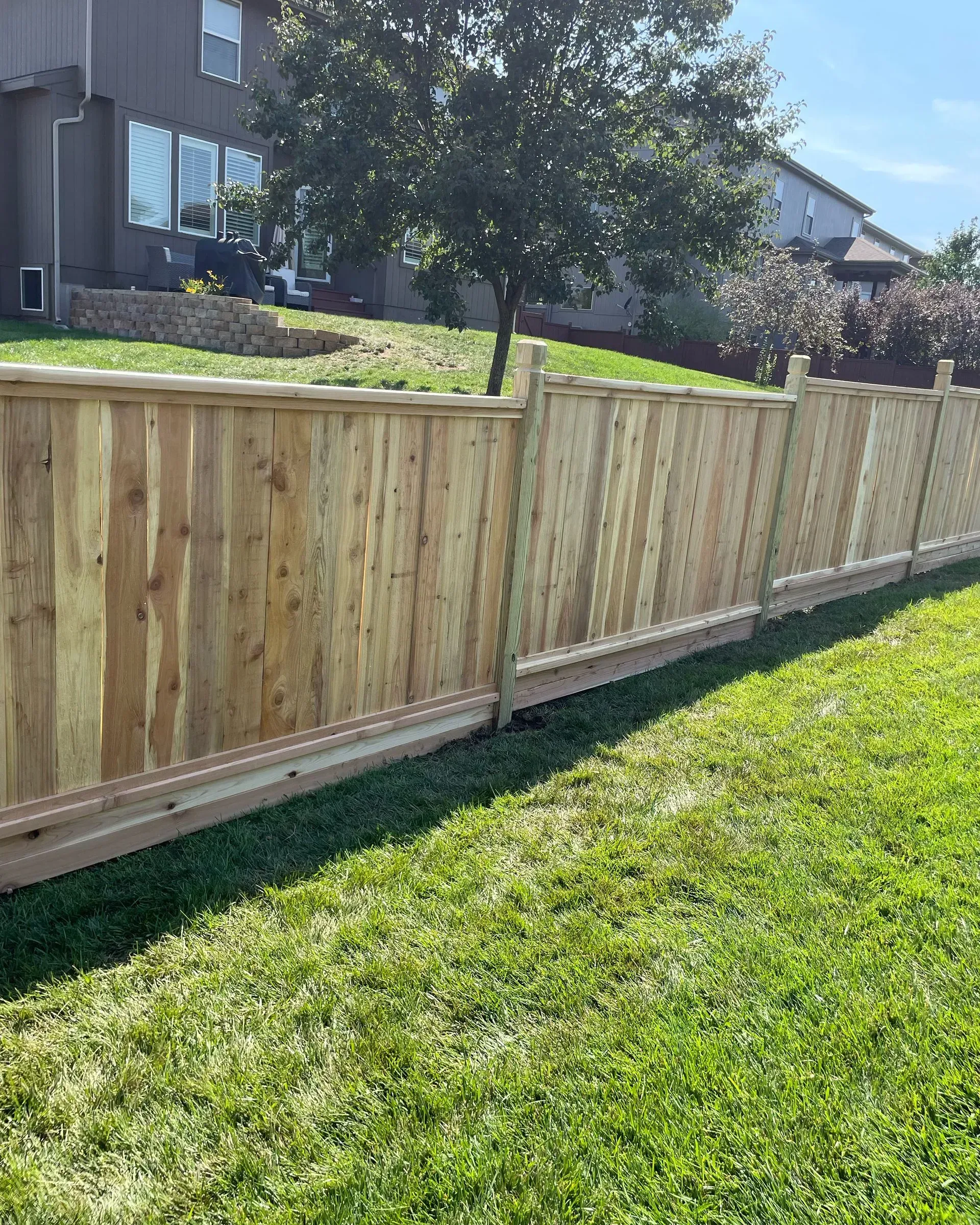 Wooden fence in a grassy yard, partially obscuring a house and tree on a sunny day.