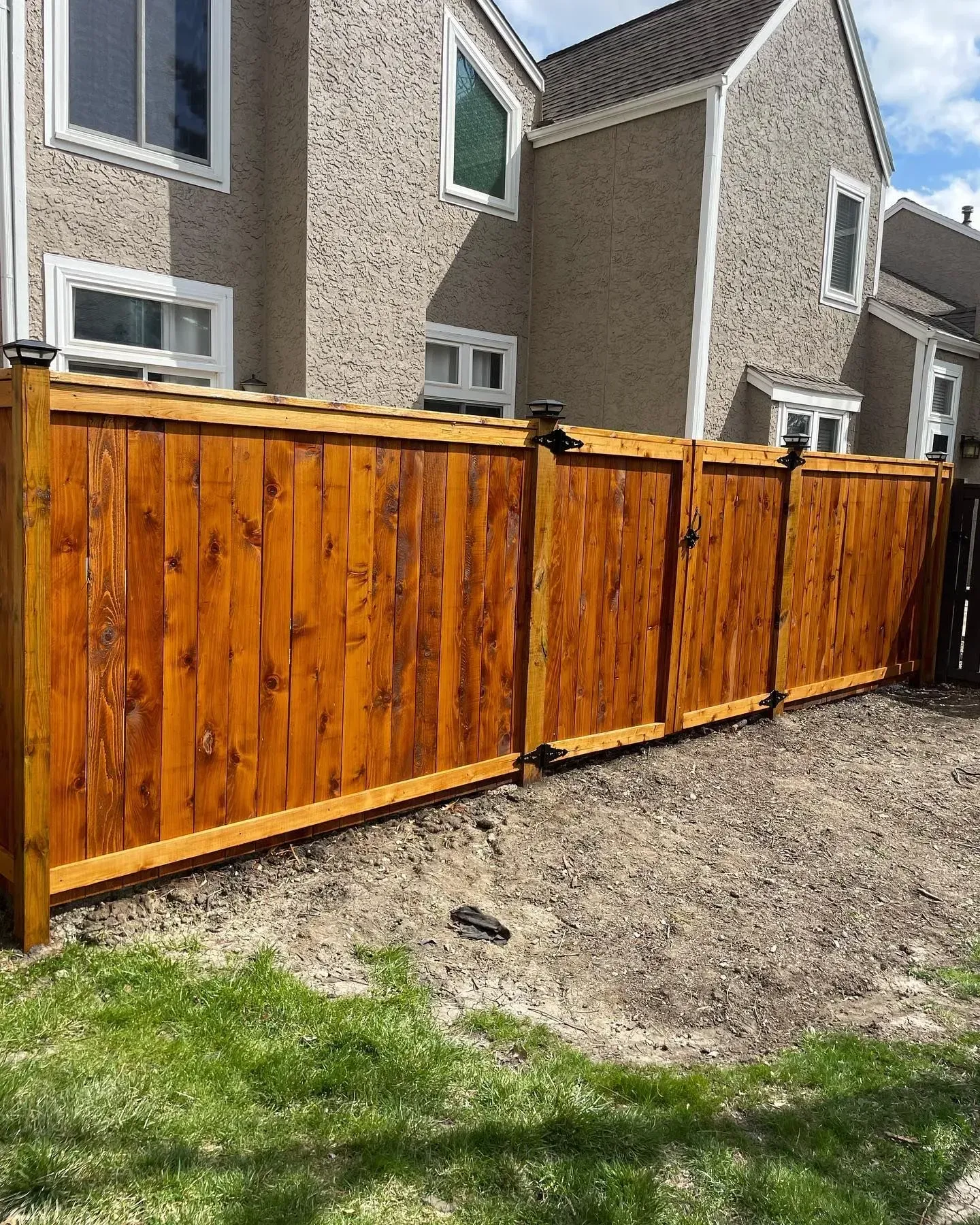 Wooden fence with a stained finish in front of a tan building with windows; a grassy lawn.