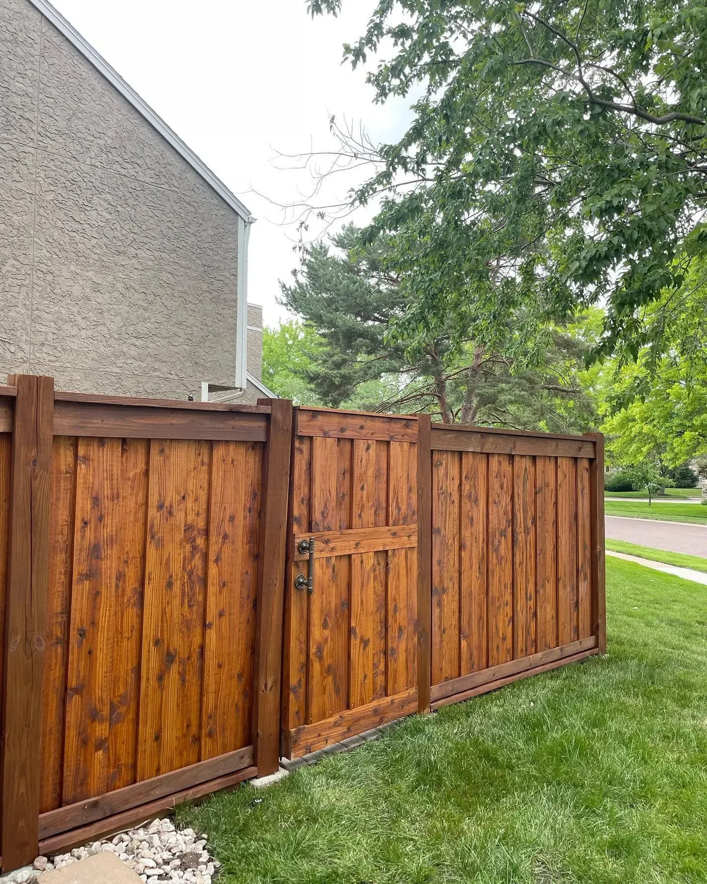 Wooden fence with gate, stained brown, in front of a tan building and green grass.