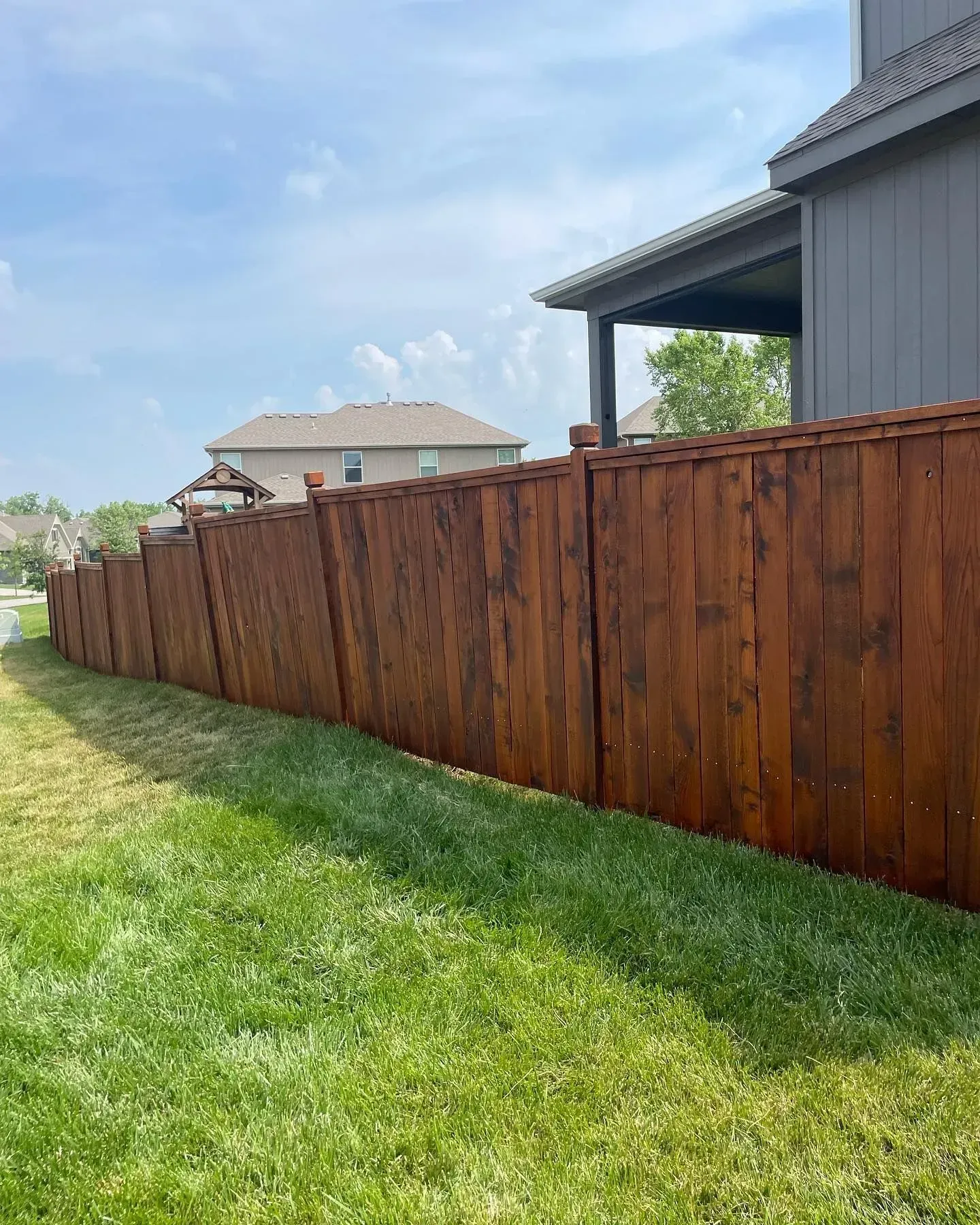 Wooden fence stained dark brown in a residential backyard, alongside green grass under a blue sky.