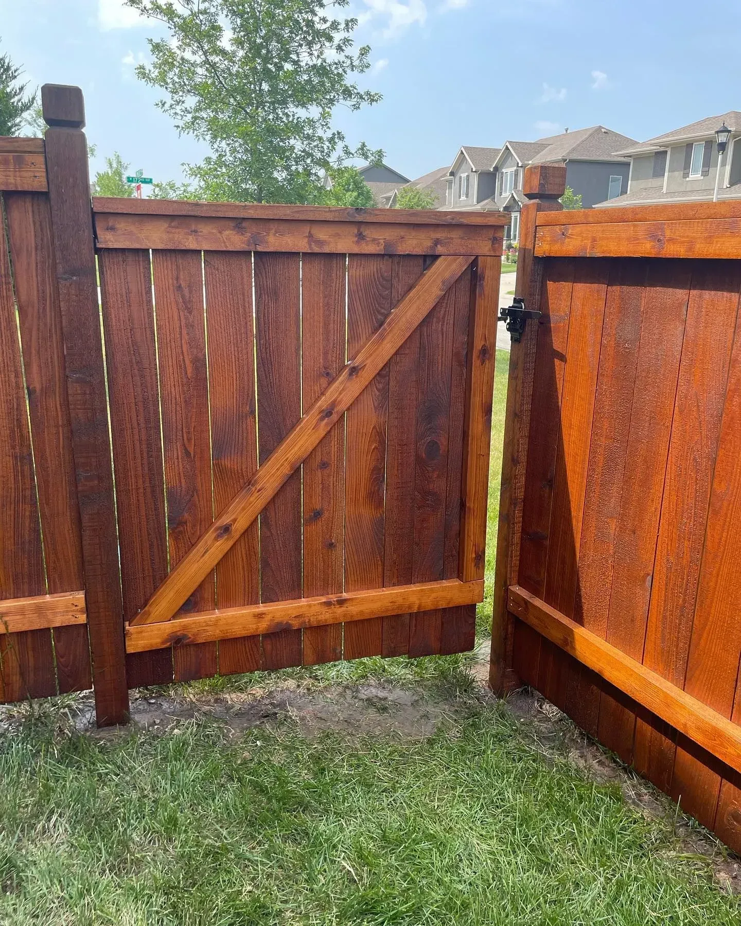Wooden gate, stained brown, open on a grassy lawn with a residential neighborhood in the background.