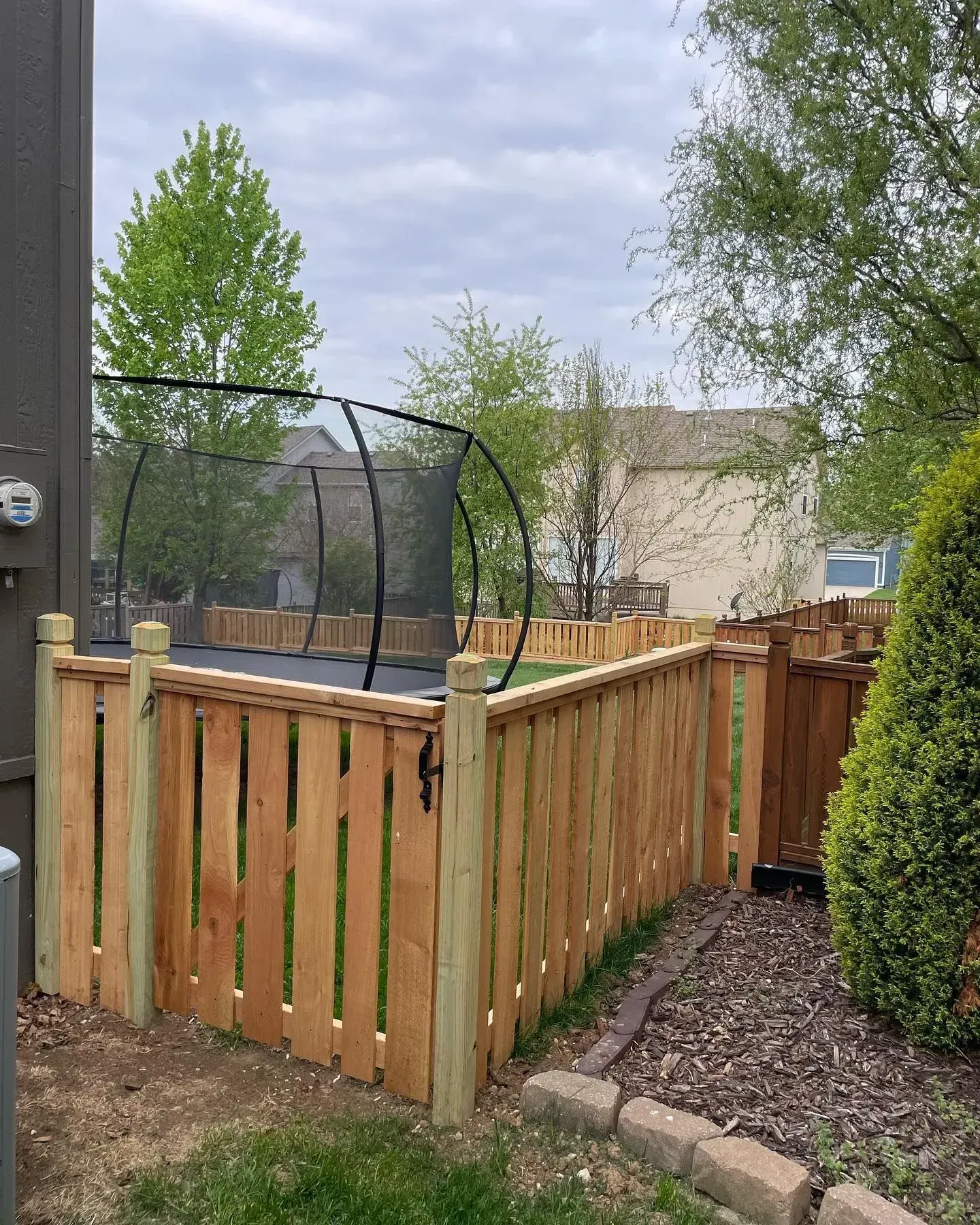 Wooden fence encloses a trampoline in a backyard. The trampoline has a black safety net.