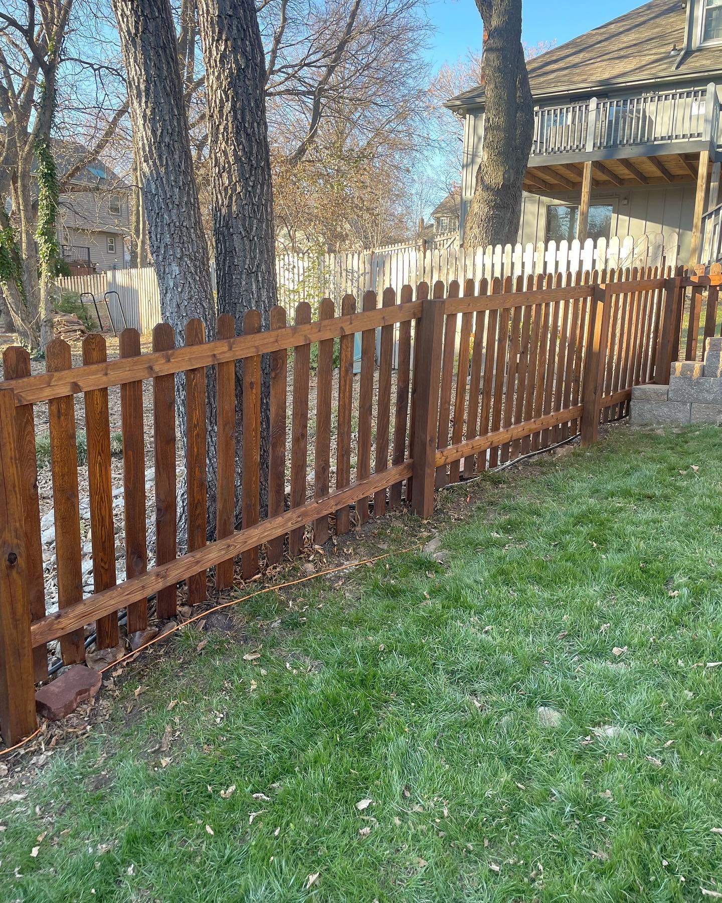 Brown picket fence in a grassy yard, with trees and a house in the background.