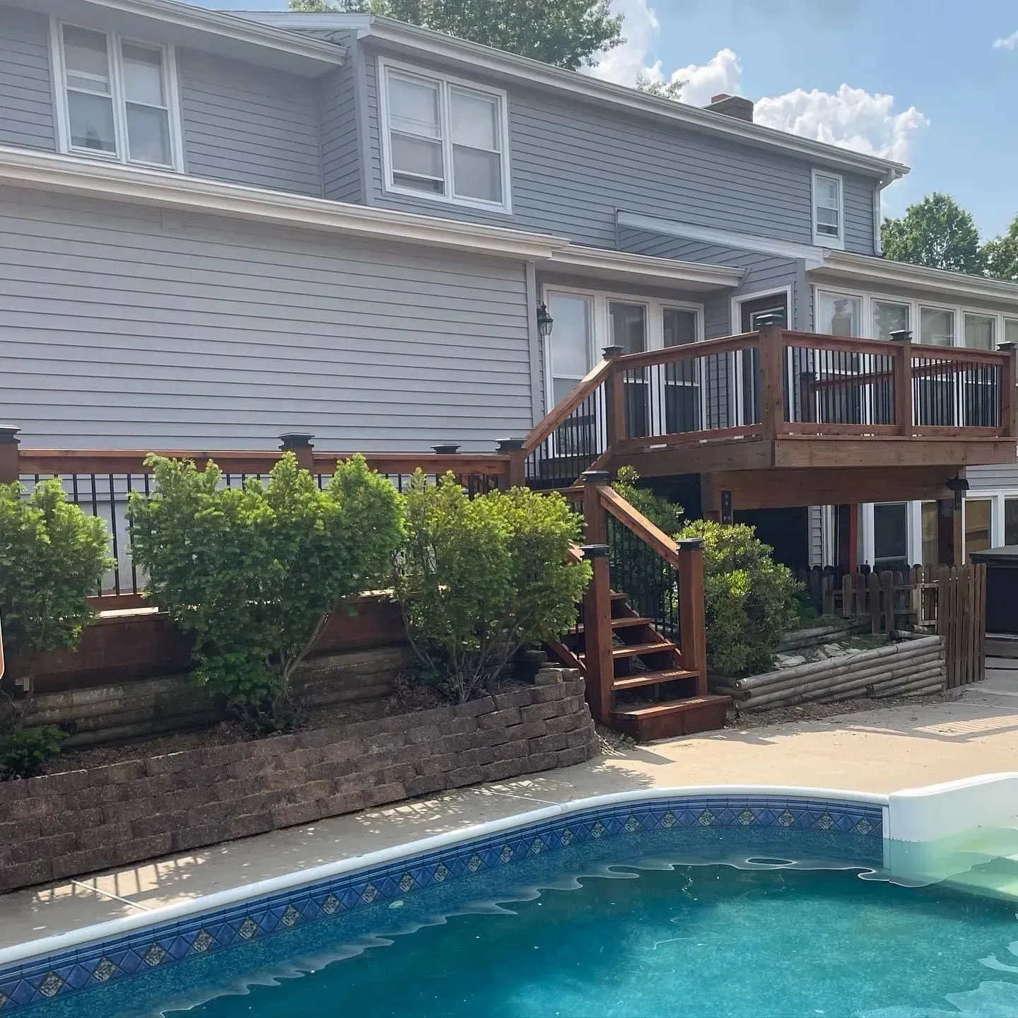 Two-story gray house with a wooden deck and pool. Lush green bushes line the deck. Blue pool.