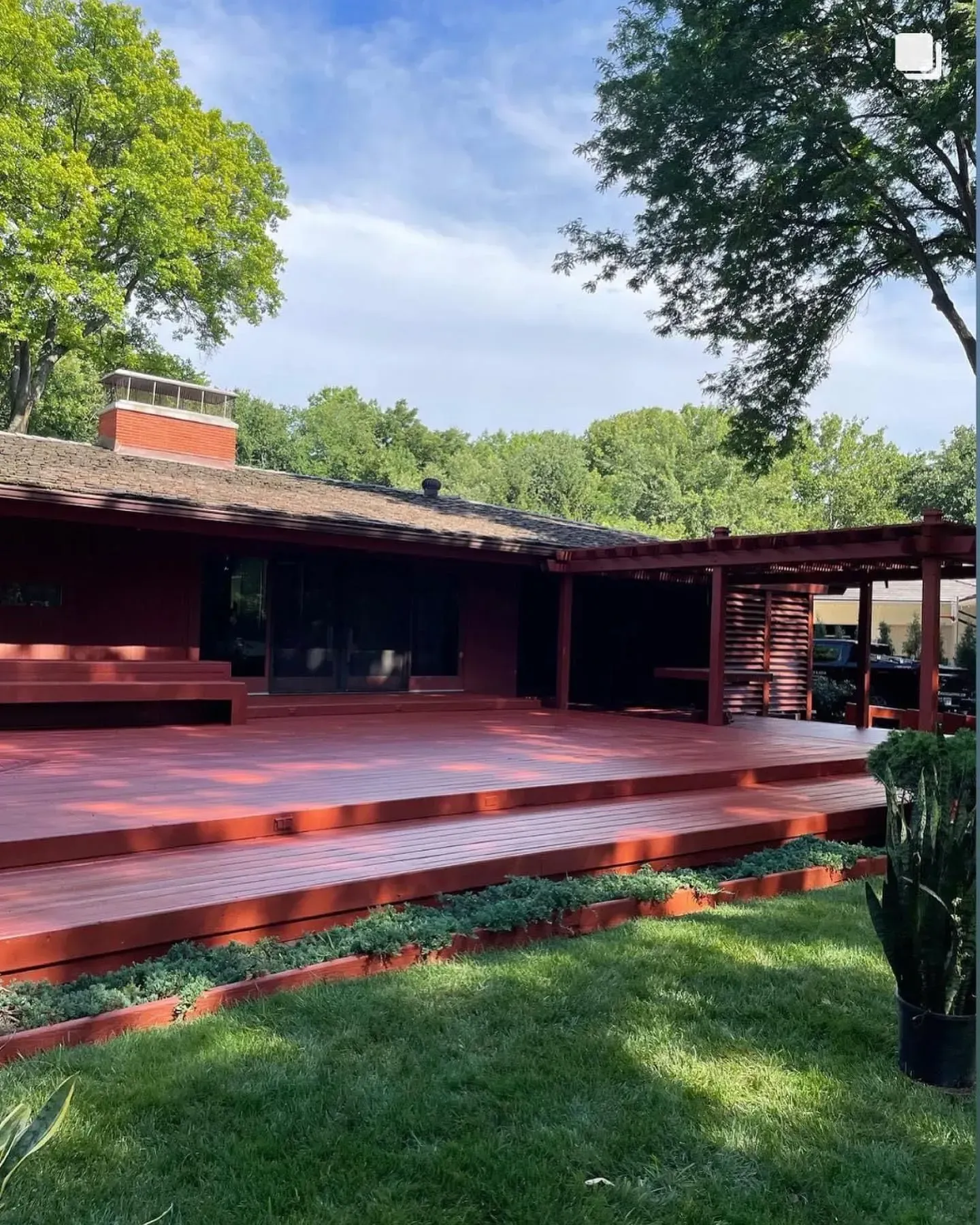 Red-painted deck with tiered levels, leading to a dark red house with a pergola, greenery, and trees.