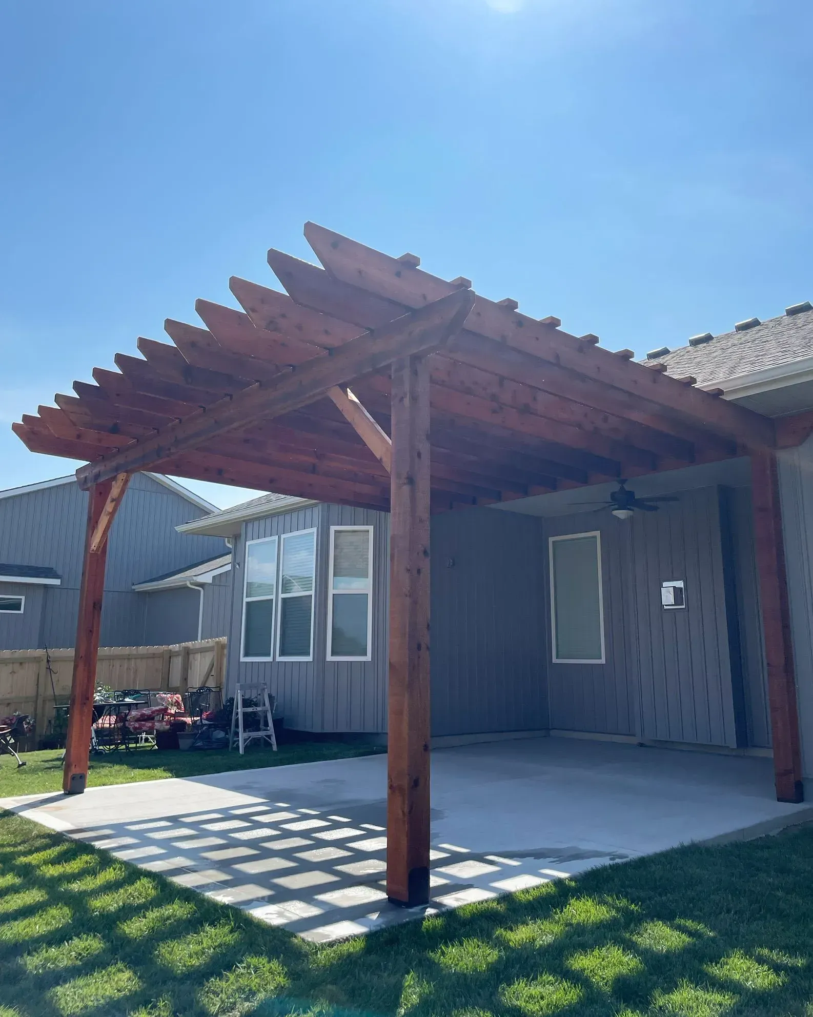 Wooden pergola over a concrete patio; built against a gray house. Sunny, exterior shot.