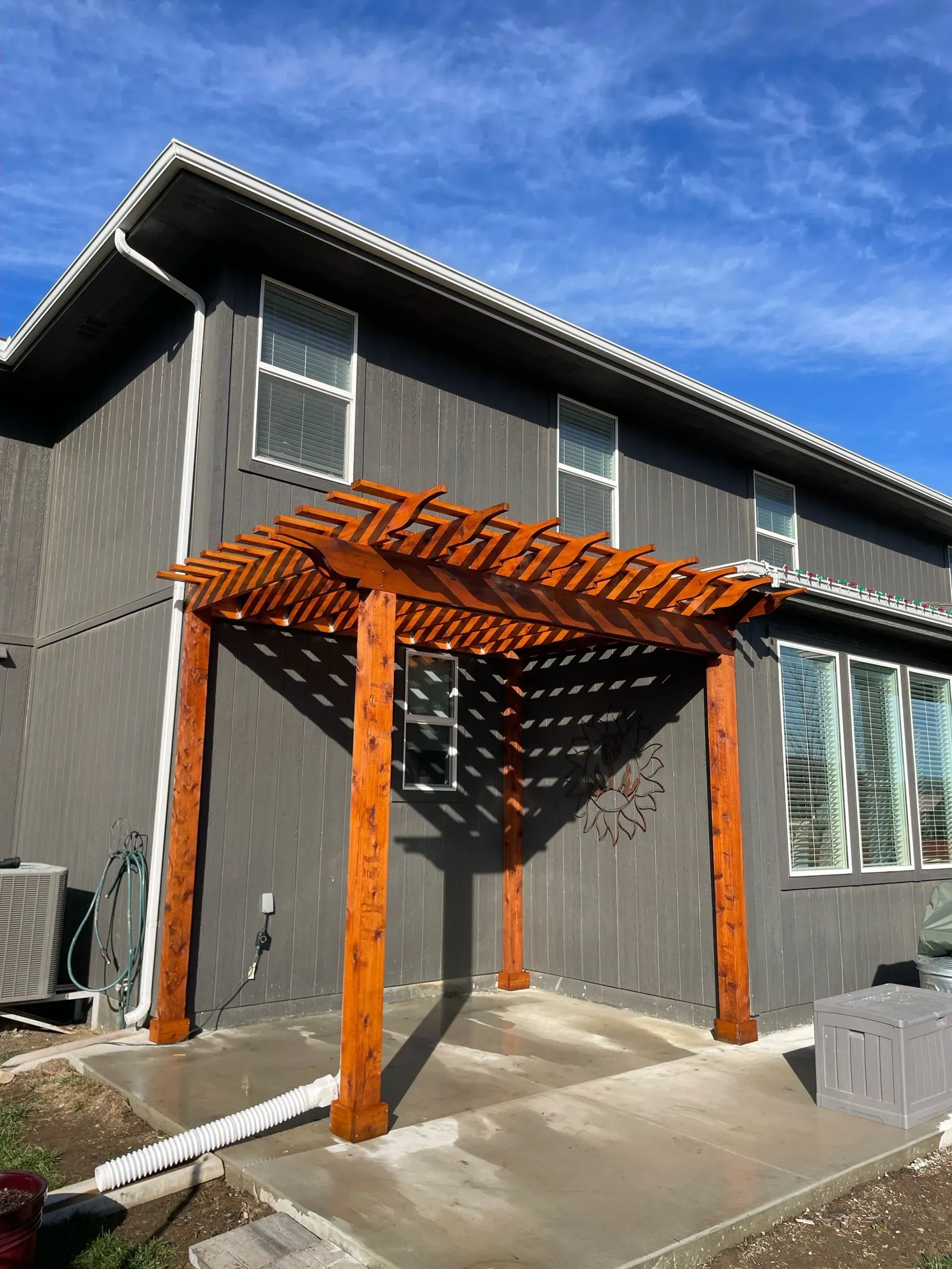 Wooden pergola attached to a gray house, casting shadows on a concrete patio under a blue sky.