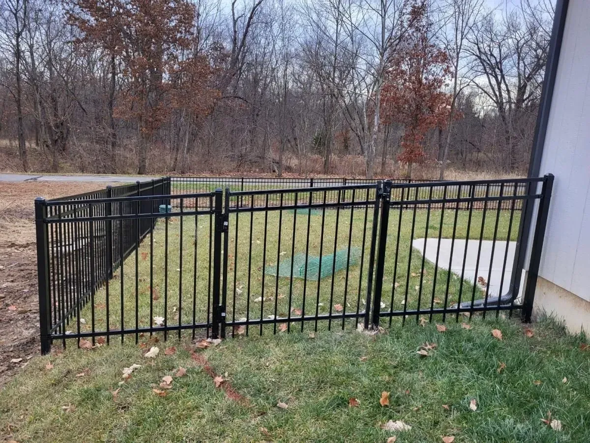 Black metal fence surrounds a grassy area next to a building. Trees in the background.