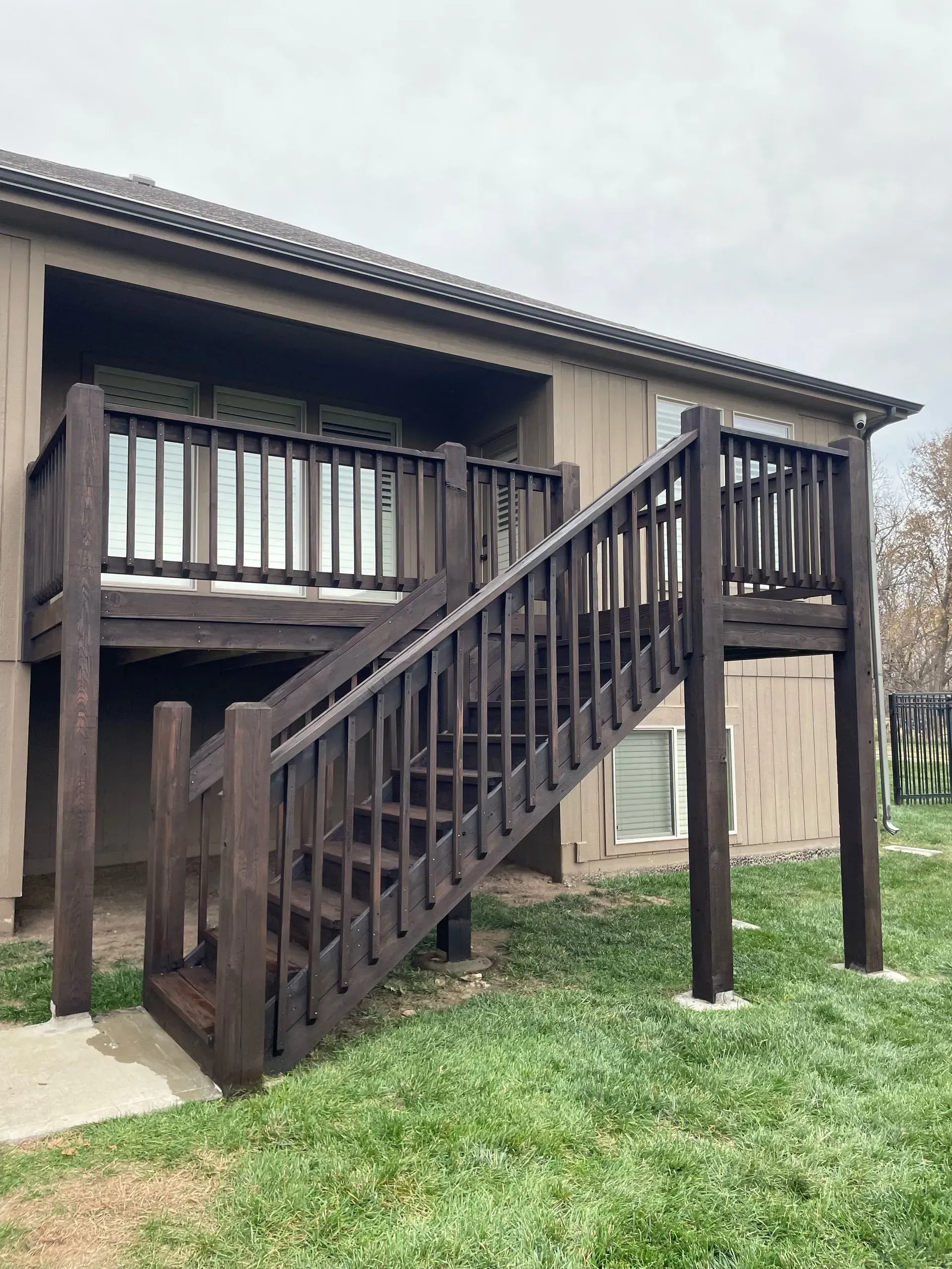A brown wooden deck with stairs leading up to the second story of a brown building, set in grassy yard.