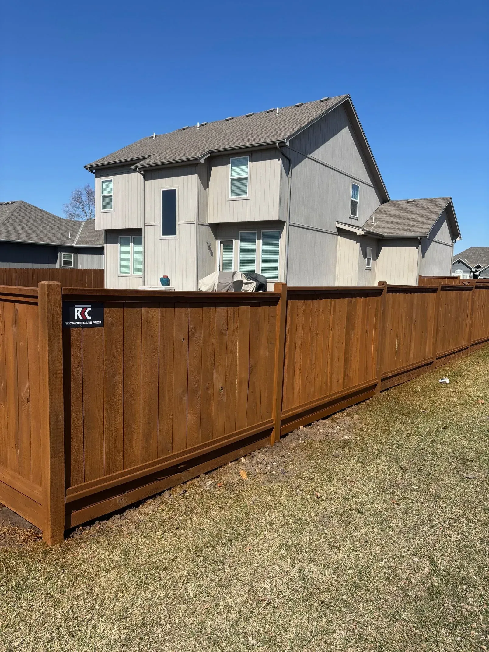 Brown wooden fence in front of a two-story house with a stucco exterior under a blue sky.