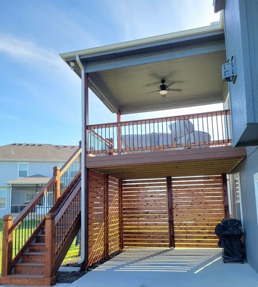 Two-story deck with wooden stairs, railing, and horizontal privacy screens. Upper level has a ceiling fan and covered seating.
