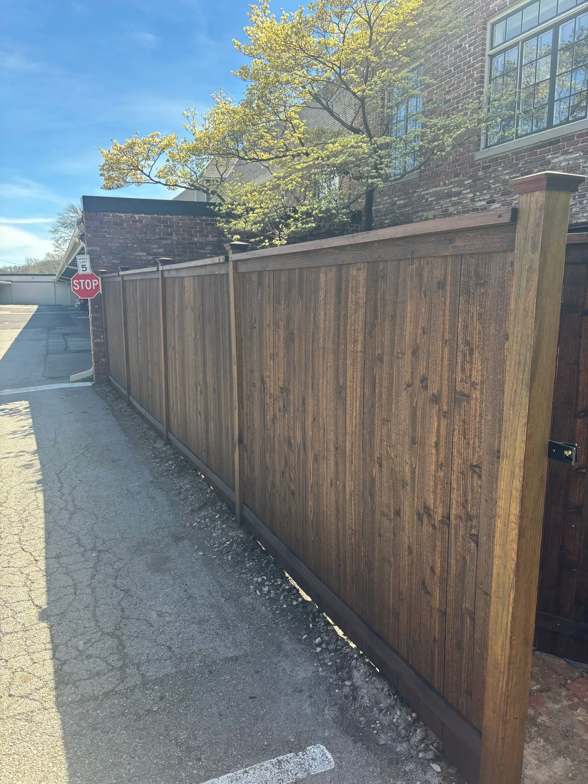 Wooden fence along a gravel alleyway, with a stop sign and brick building visible in the background.