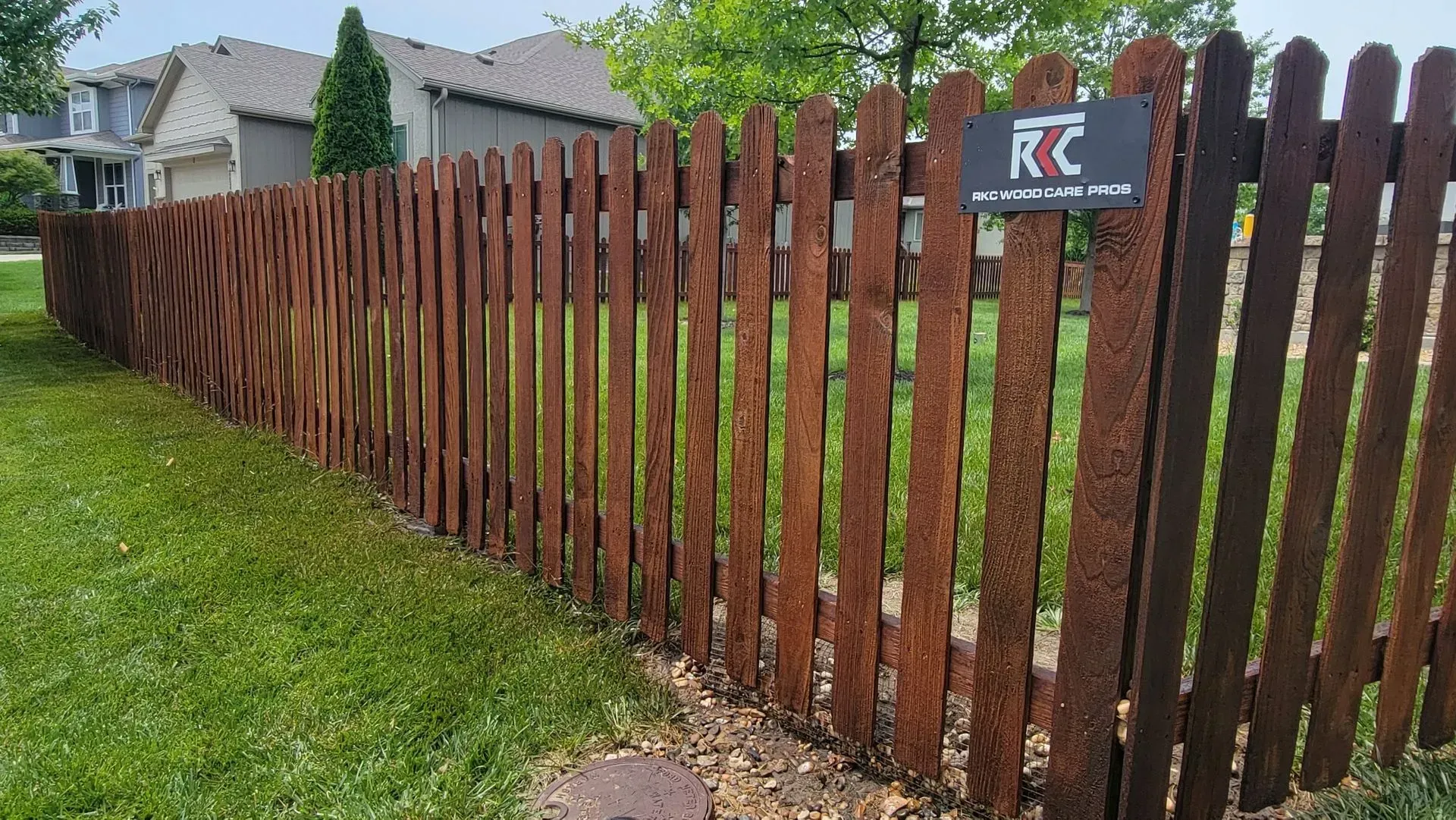 Brown, rusty-looking metal picket fence in front of green grass and houses.