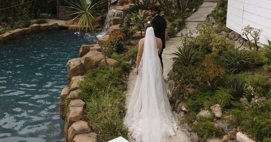 Bride and groom walking on a pathway beside a pool, surrounded by greenery. Wedding attire. Outdoor setting.