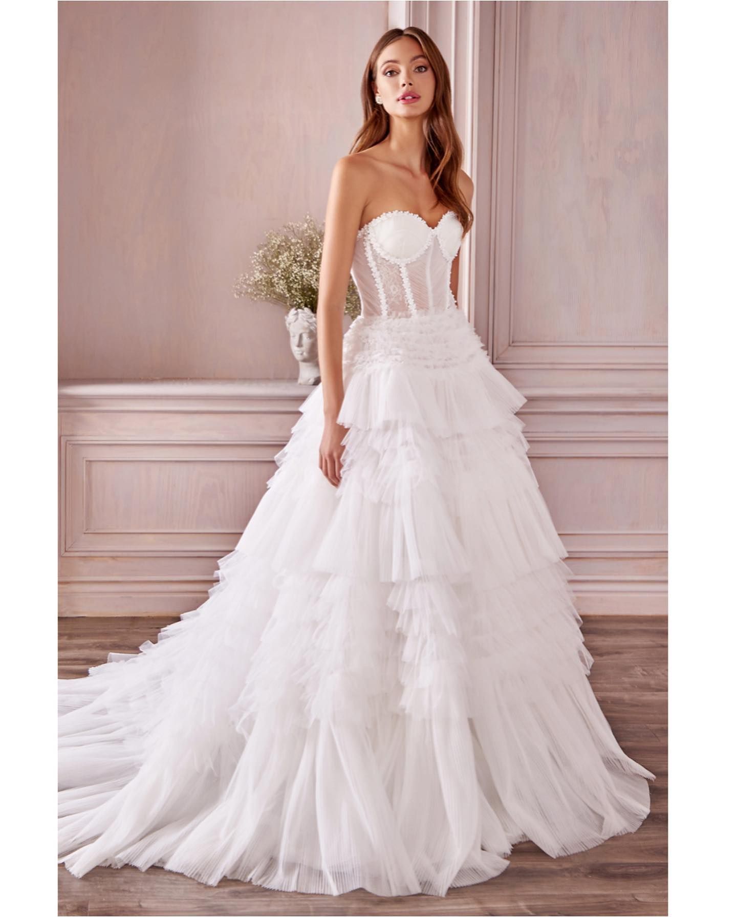 Woman in a white strapless wedding dress with a tiered skirt, posing in a room with a light pink wall.