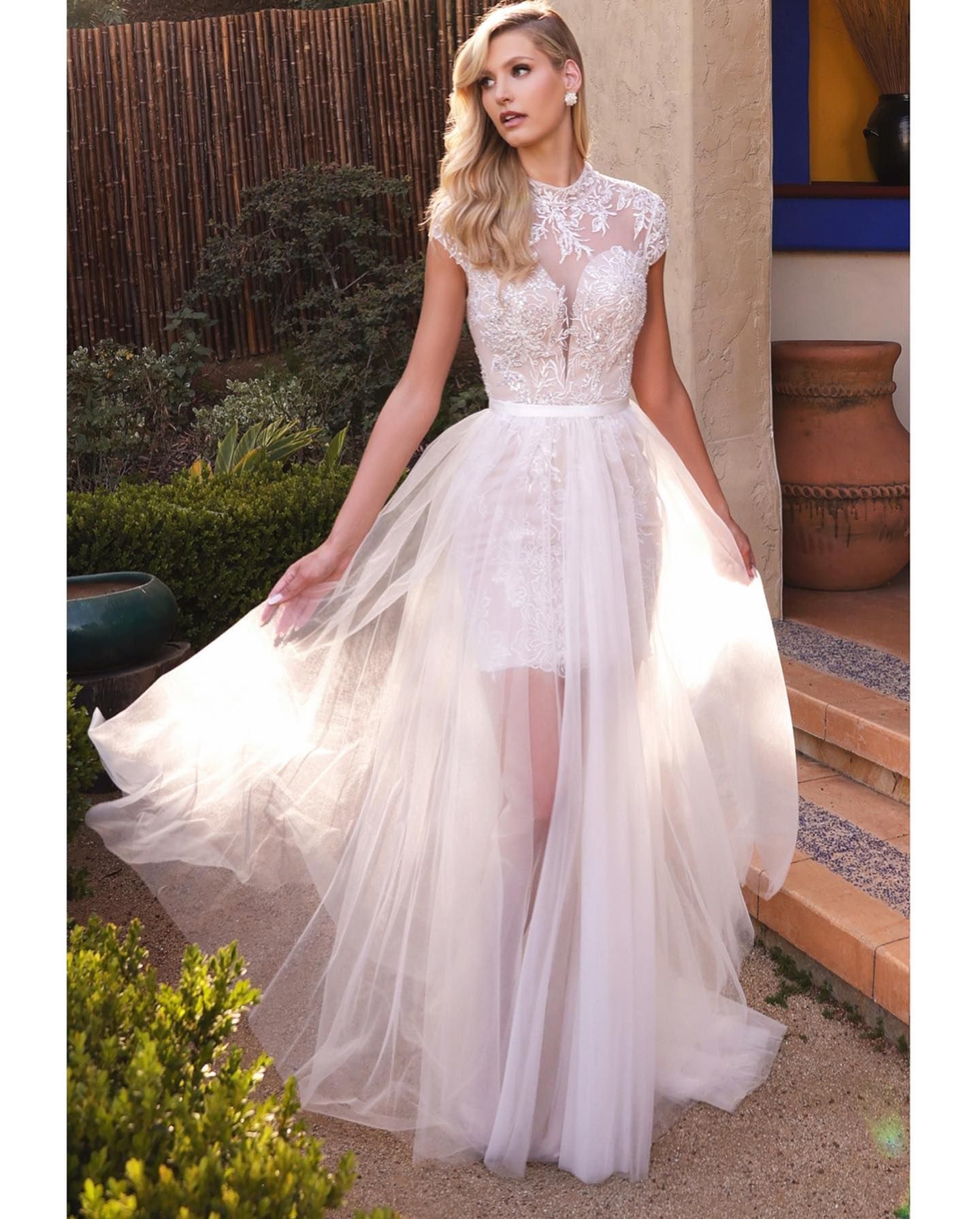 Woman in white wedding dress with flowing tulle skirt, standing outdoors near plants.
