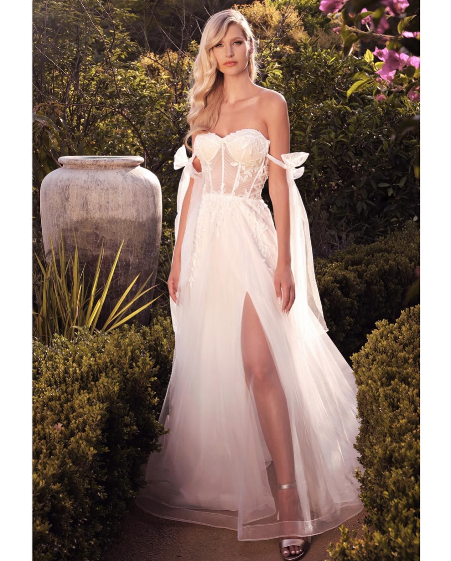 Woman in white strapless gown with bows, high slit, standing in a garden with greenery and a stone vase.