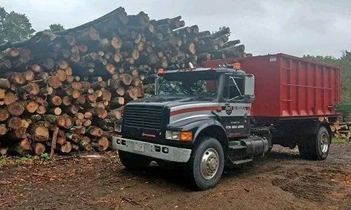 Black and red dump truck loaded with logs in front of a large wood pile.