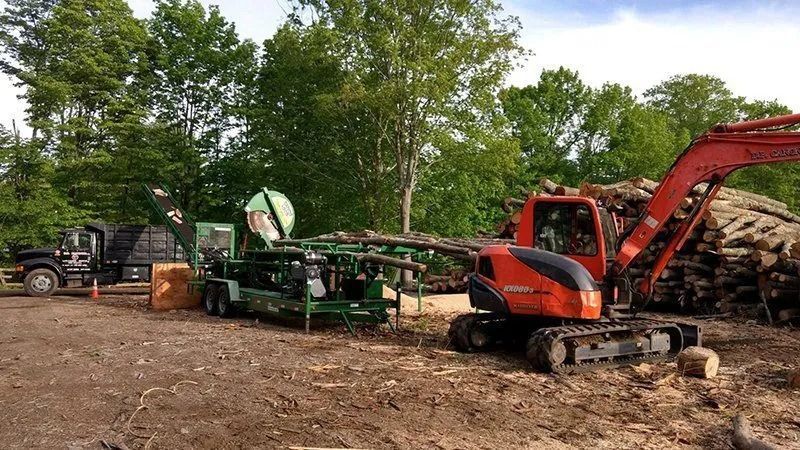 A wood processing operation with an excavator, a wood chipper, and logs.