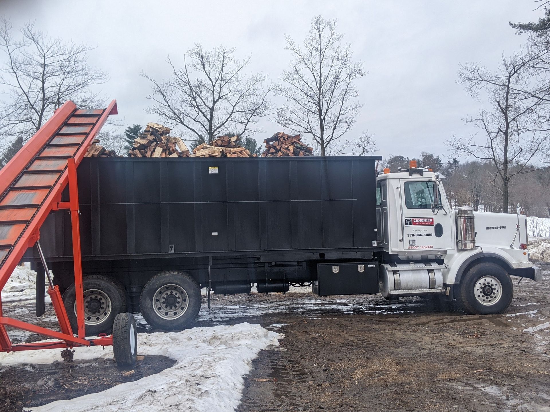 White truck with black container filled with firewood, near an orange conveyor belt, in a snowy setting.
