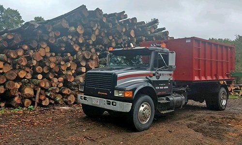 Truck loaded with red container beside a large pile of logs, on a dirt surface.