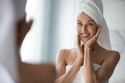 Woman in a towel, looking in mirror, smiling and touching face. Bathroom setting.