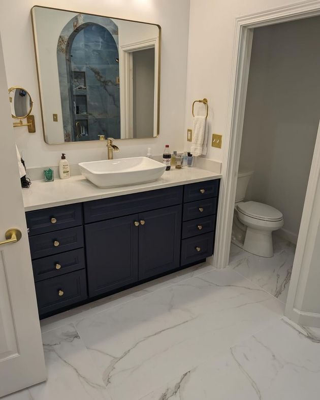 Bathroom with navy vanity, white countertop, gold fixtures, large mirror, and adjacent toilet room.