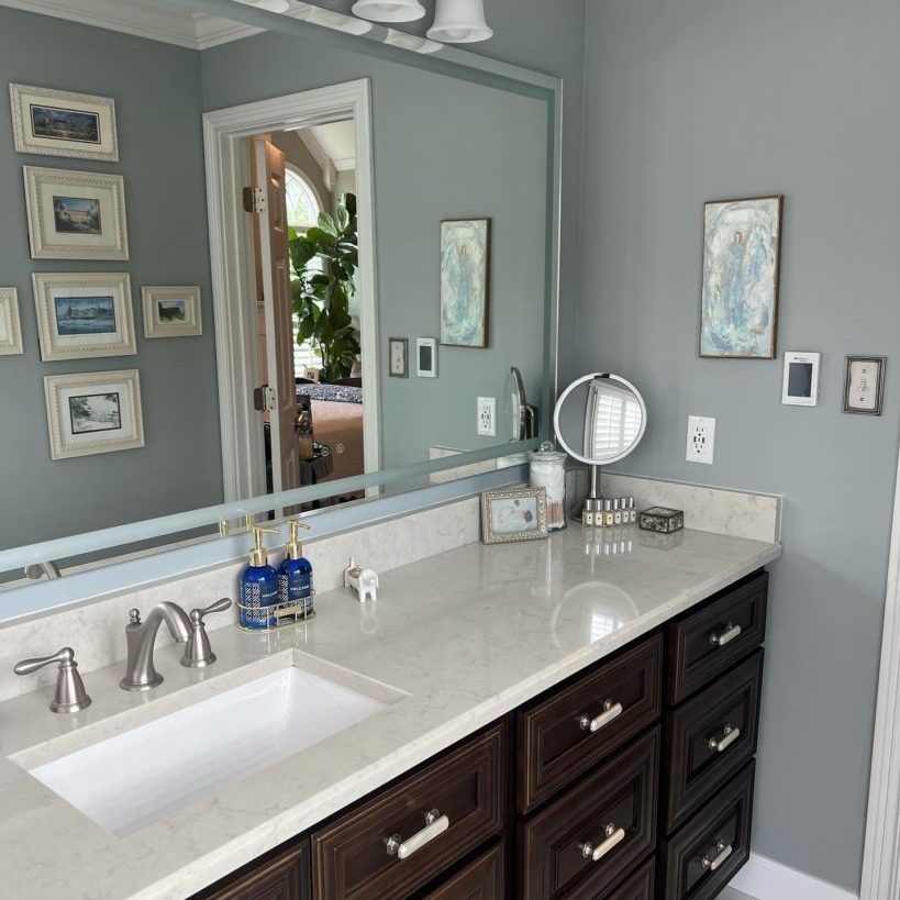 Bathroom with a long mirror, countertop, dark brown cabinets, and blue-gray walls; decorated with art.