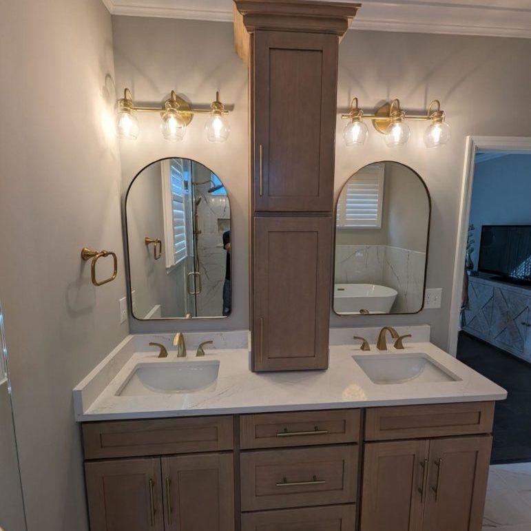 Bathroom with dual sinks, arched mirrors, and a tall cabinet. Light wood cabinetry, gold fixtures.