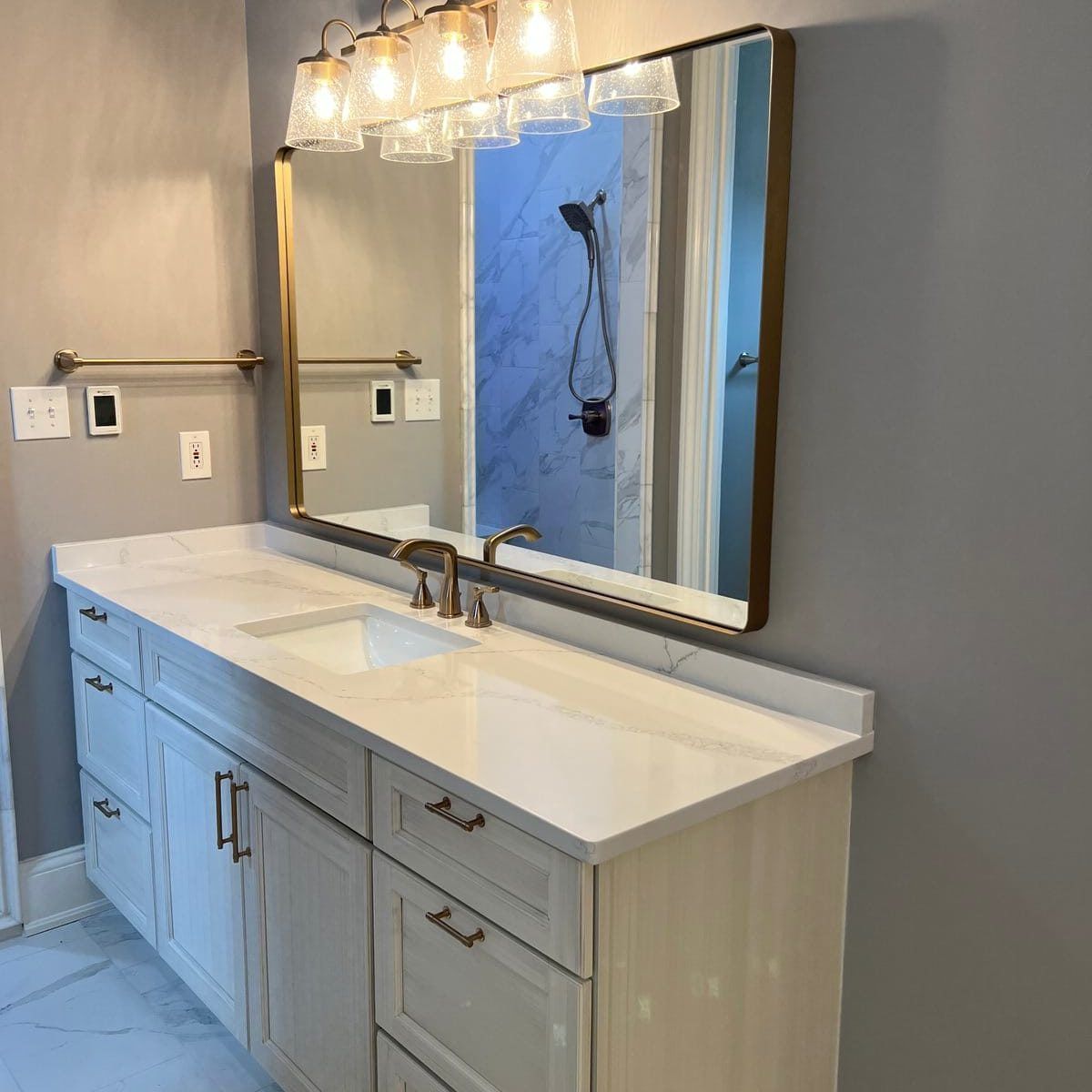 Bathroom vanity with white countertop, cabinets, large mirror, and gold fixtures.