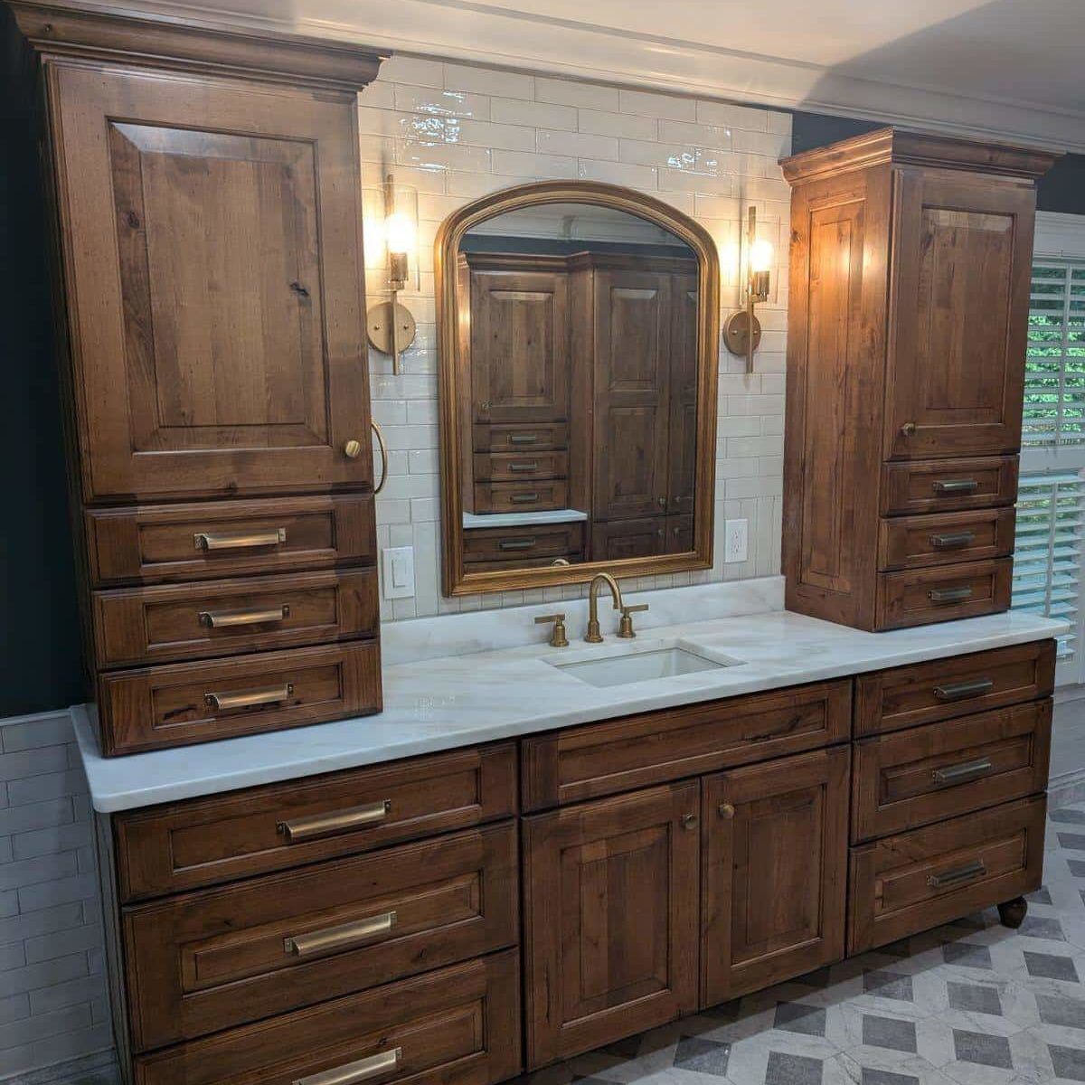 Bathroom with wooden cabinets, marble countertop, gold mirror and fixtures, and decorative tile.