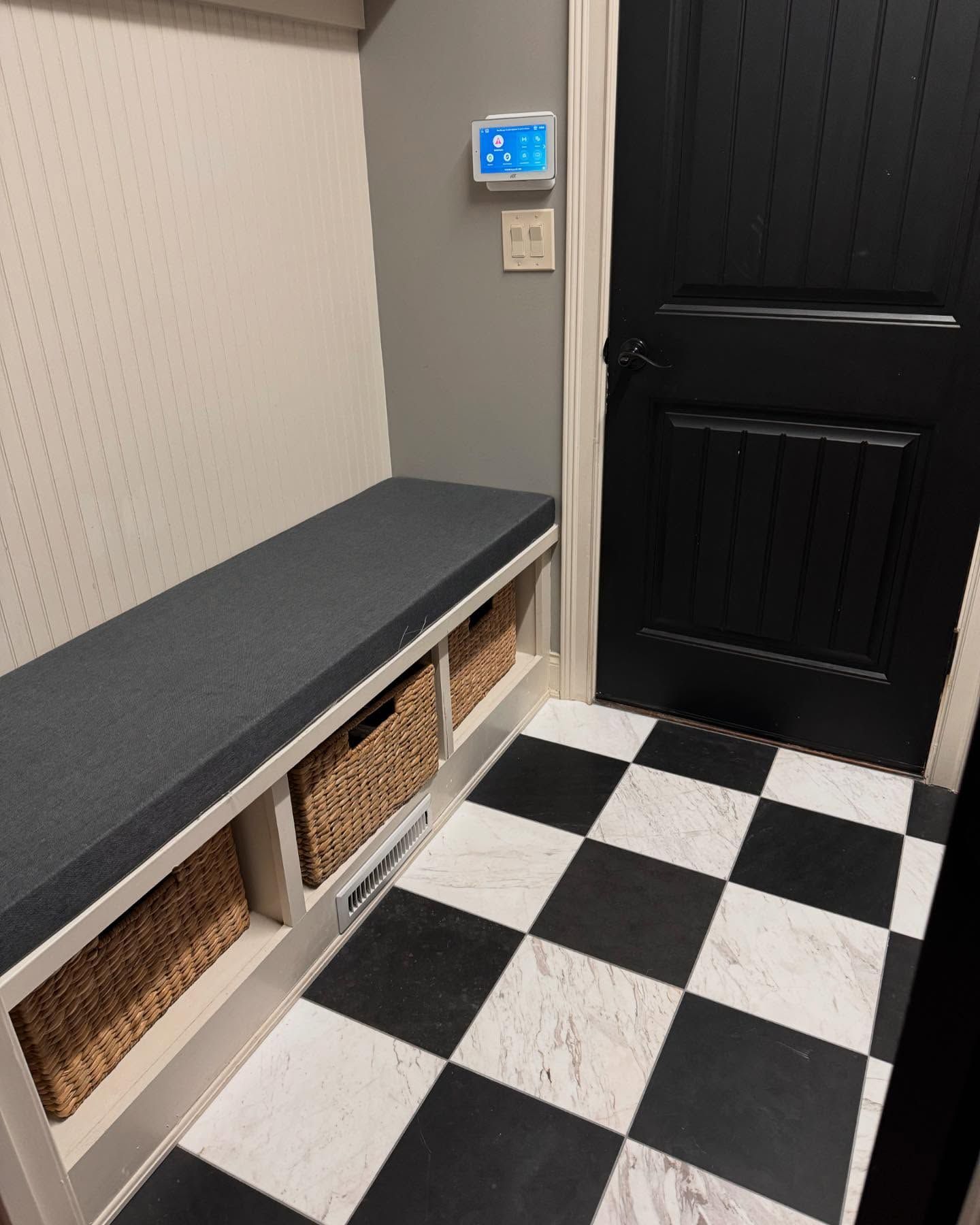 A built-in bench with storage baskets and a cushion in a room with checkered flooring, next to a black door.