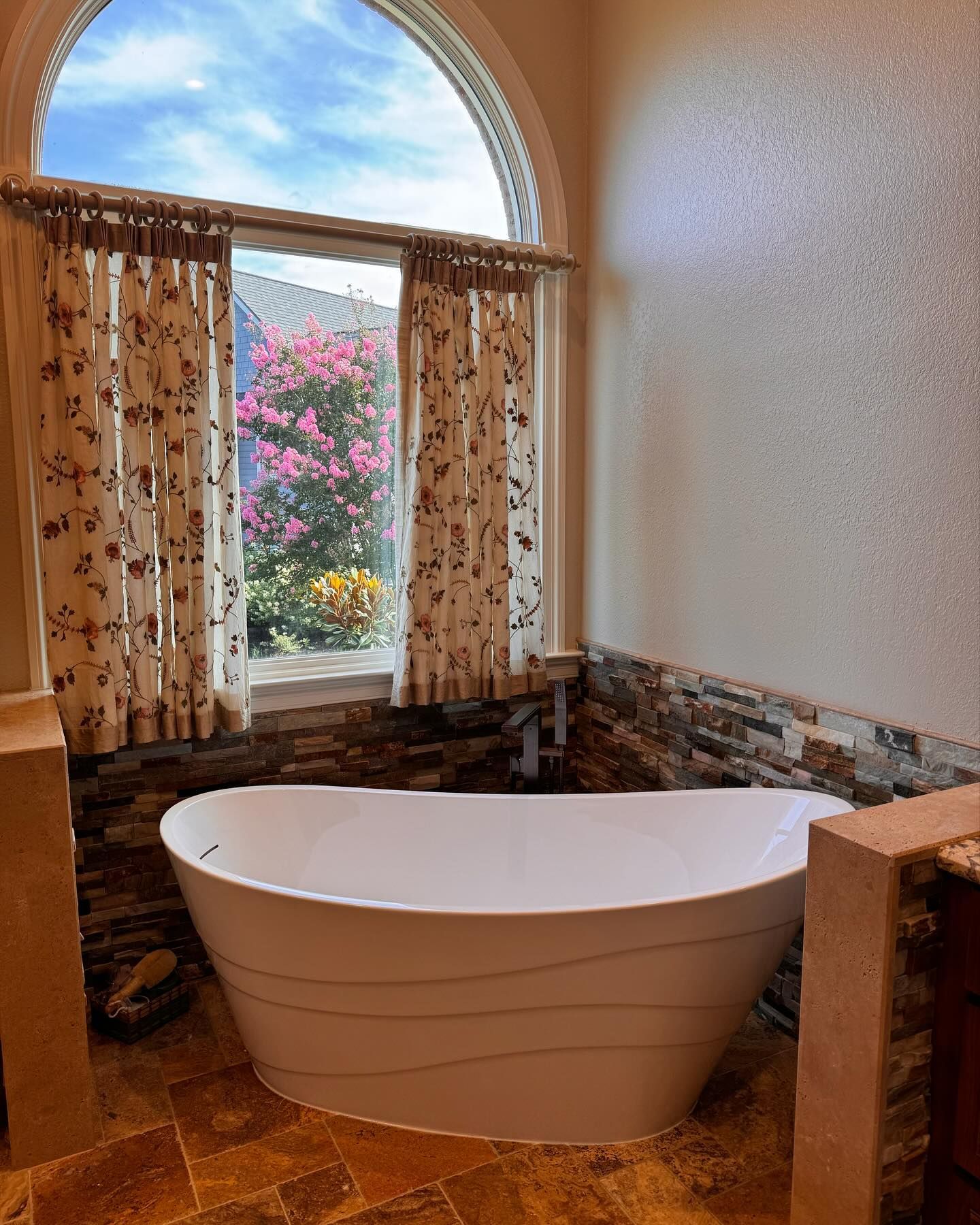A white bathtub sits beneath an arched window with floral curtains, stone accents, and a view of pink flowers.
