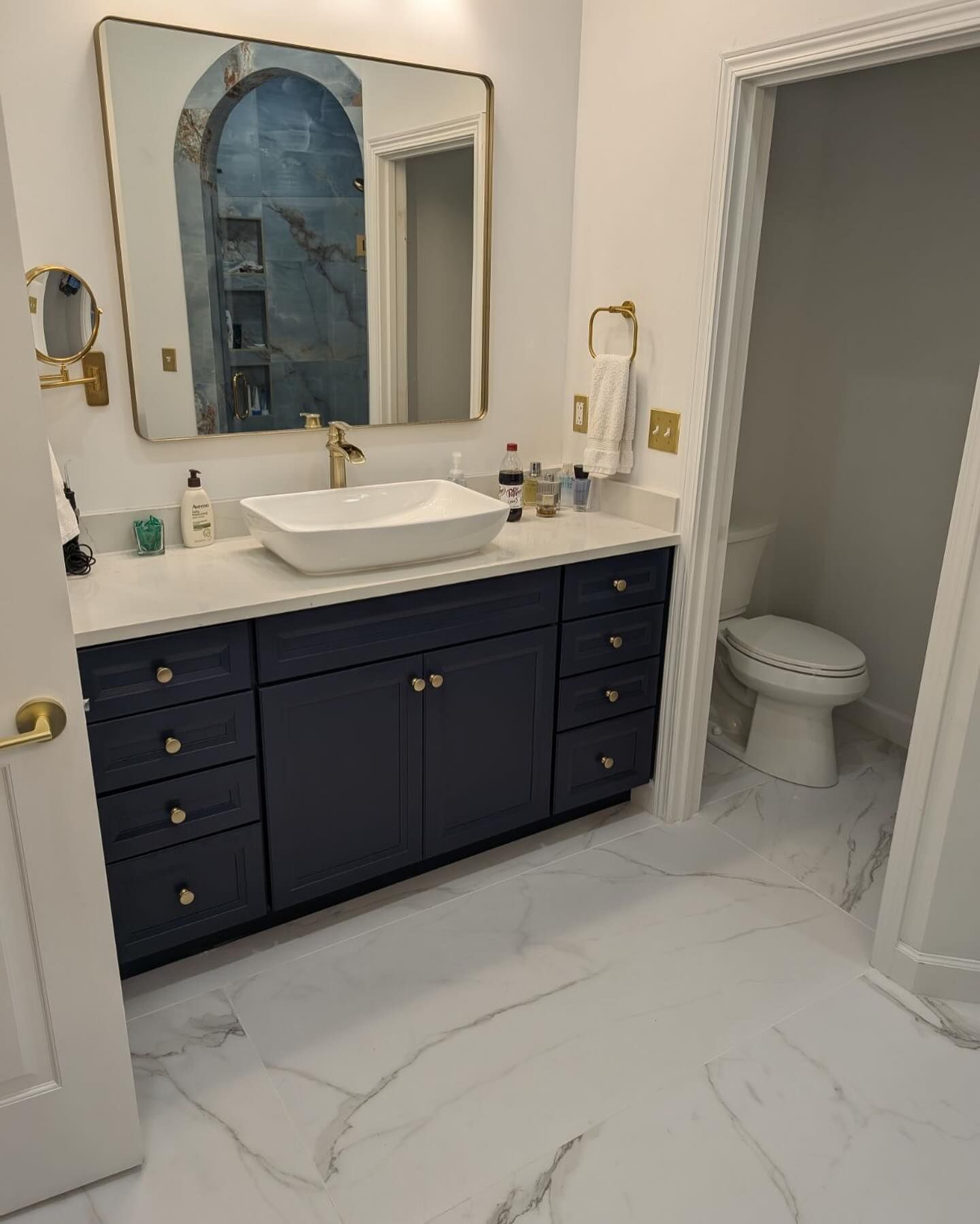 Modern bathroom with blue vanity, gold fixtures, marble-look floor and separate toilet room.
