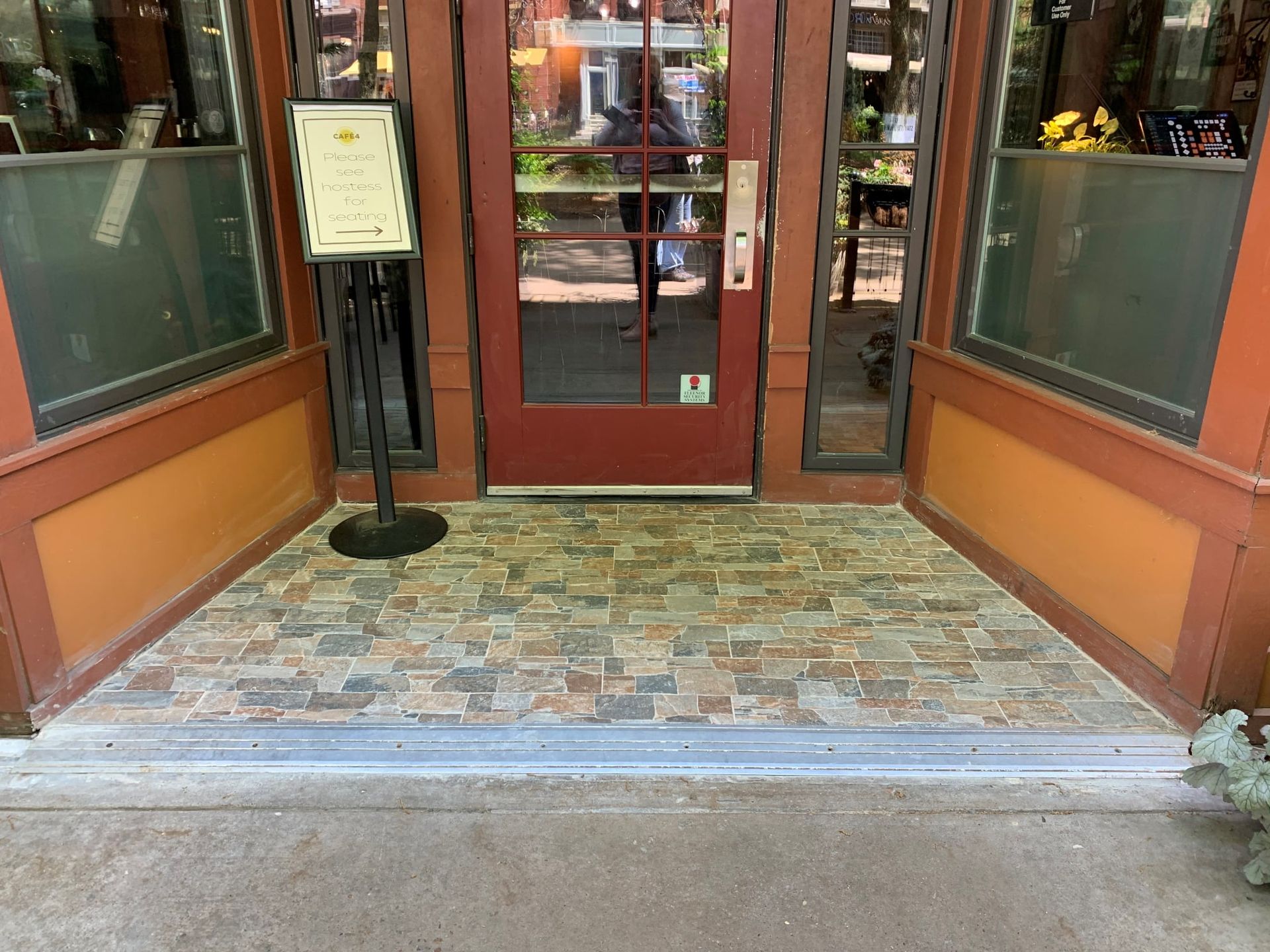 Entrance to a building with a red door, stone floor, and sign on a stand. Orange and green accents are visible.