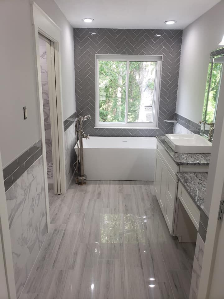 Modern bathroom with gray tiled walls and flooring, white tub, vanity, and large window.