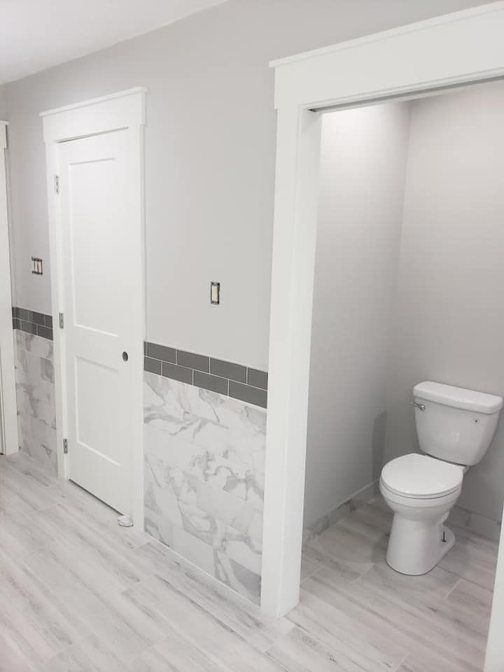 Bathroom with white toilet, door, and walls; grey tiled accent, and wood-look flooring.