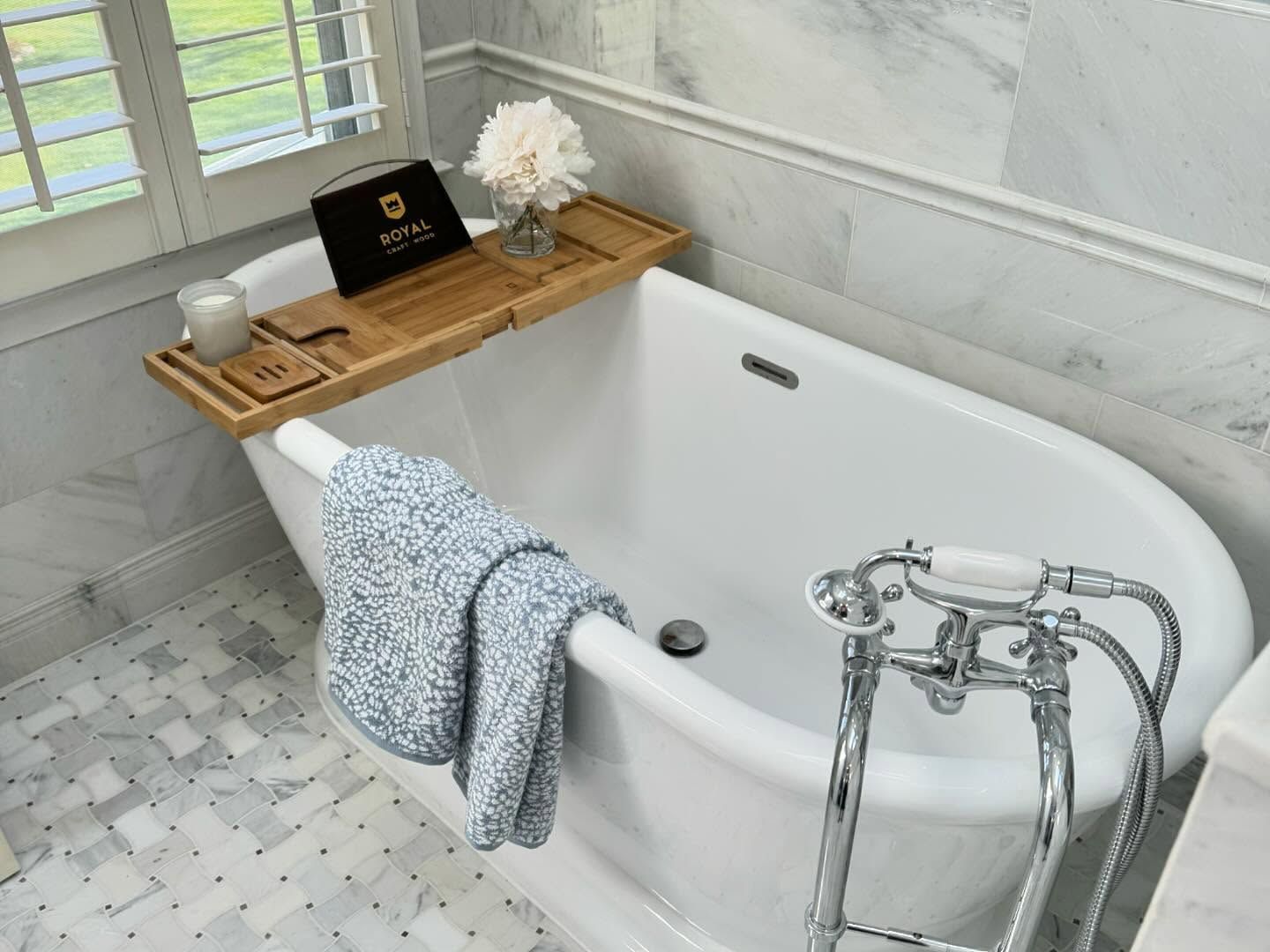 Bathtub with bamboo tray holding a book and candle, blue towel draped on the side, marble tile surround.