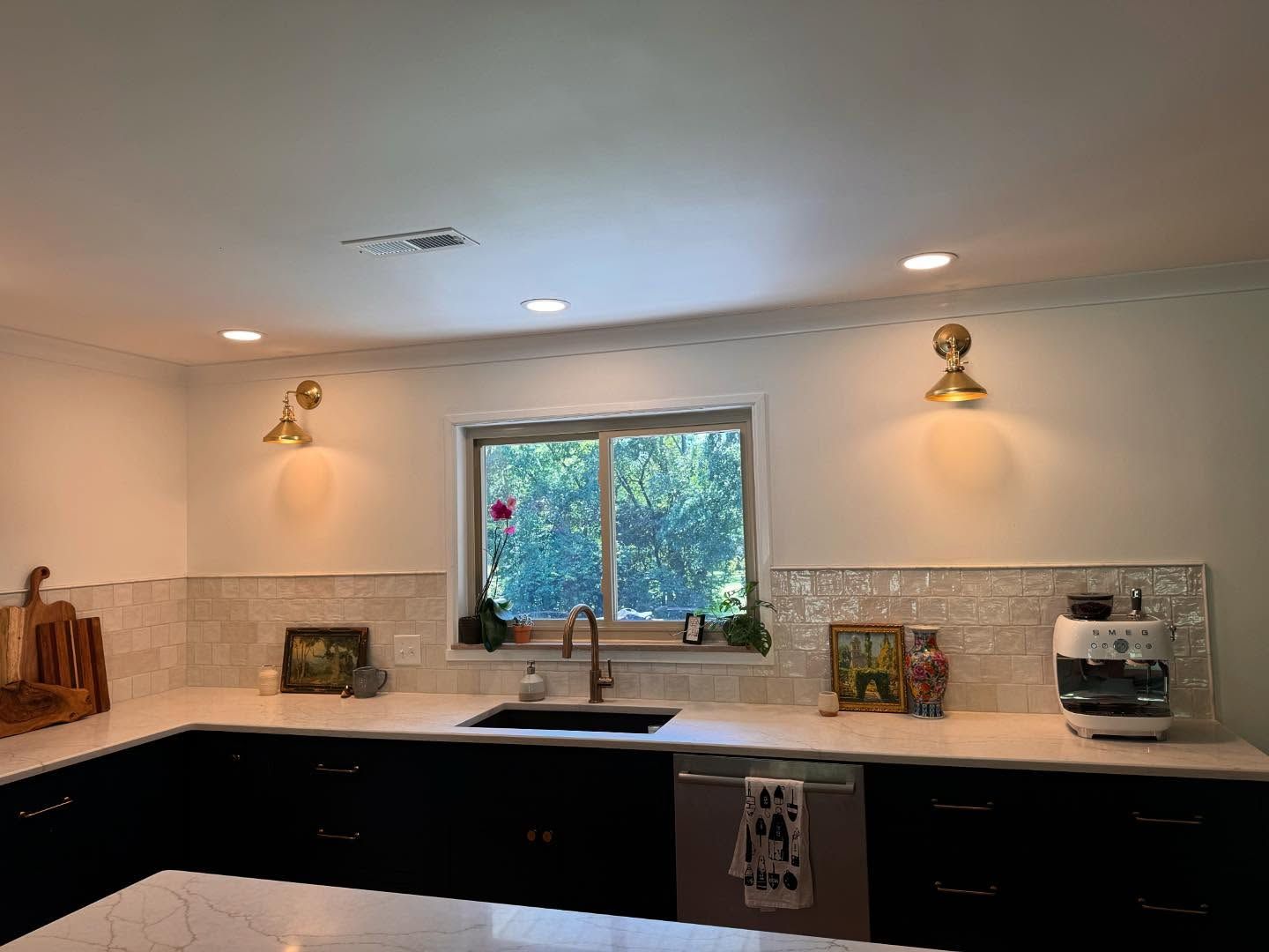 Kitchen with black cabinets, white countertops, window with sink, and brass sconces.