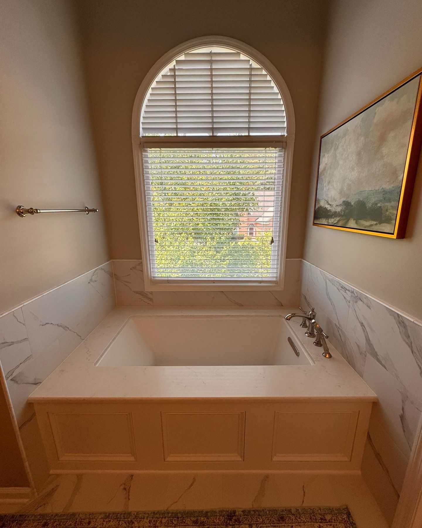 Bathroom with a built-in white bathtub, beneath an arched window with blinds, and a framed painting on the wall.